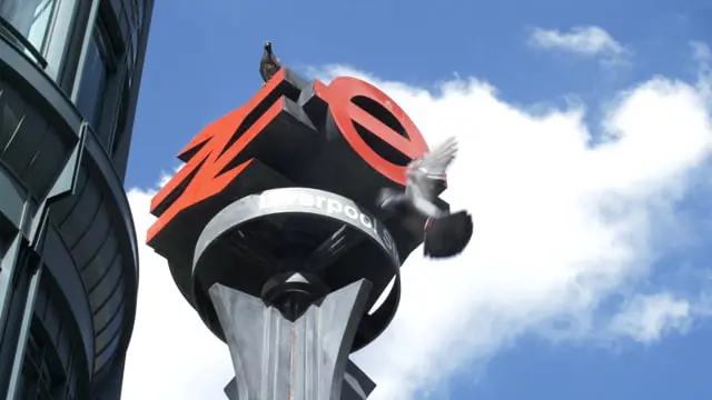 Large logo sculpture on a pole against a blue sky with clouds and a building visible