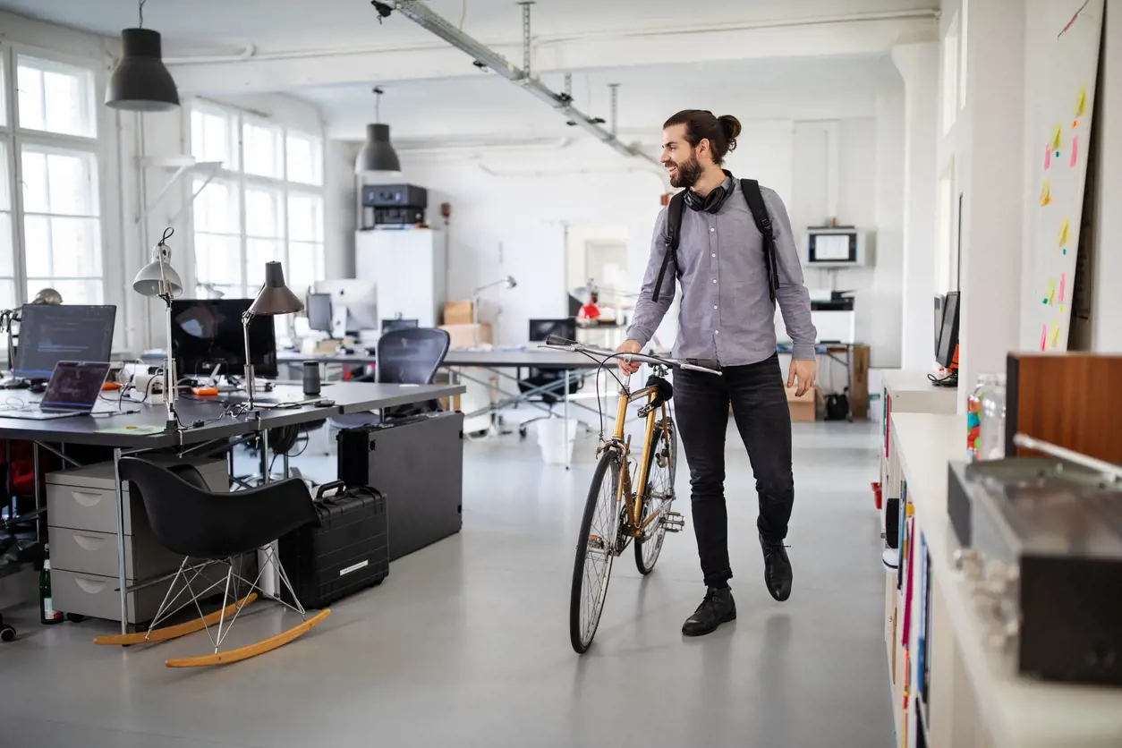 A bearded man in casual clothes walks a bicycle through an open-plan office space. 
