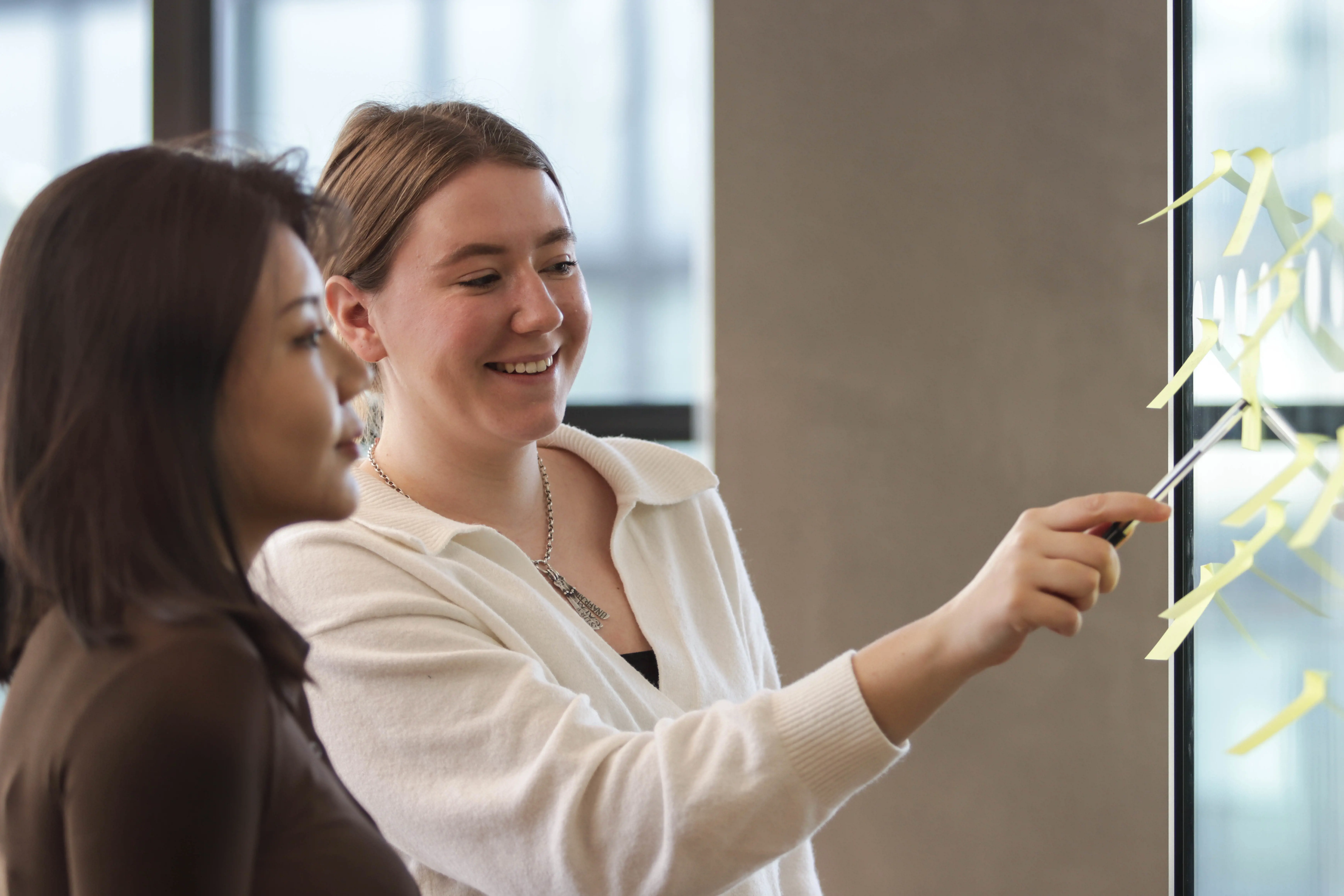 two female colleagues reviewing their brainstorming sticky notes on a glass wall 