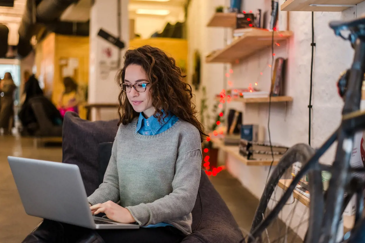 A woman with curly hair and glasses sits on a couch, working on a laptop in a casual office